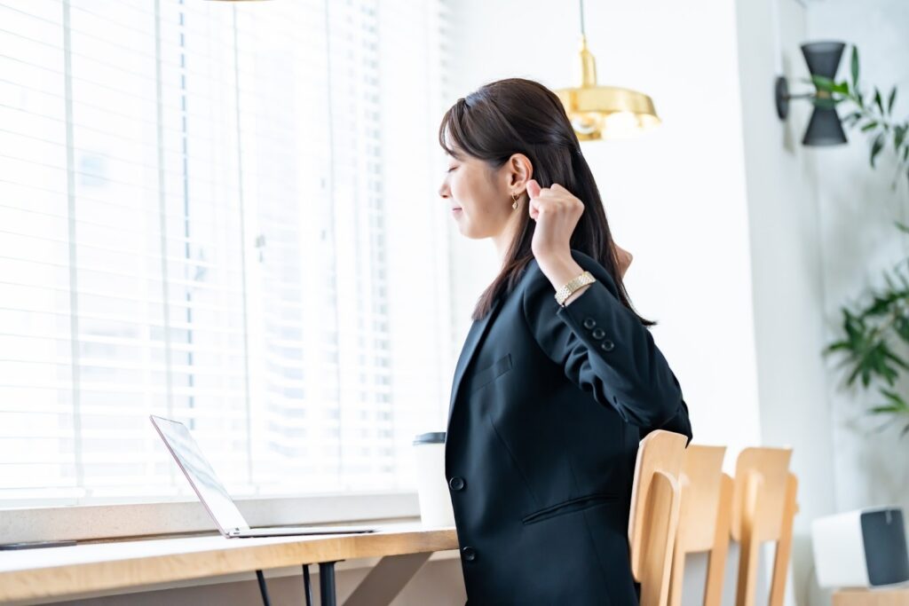 A woman in a business suit stretches and smiles while sitting by a window in a bright room. A laptop and a coffee cup are placed on the desk in front of her: Mindfulness Techniques