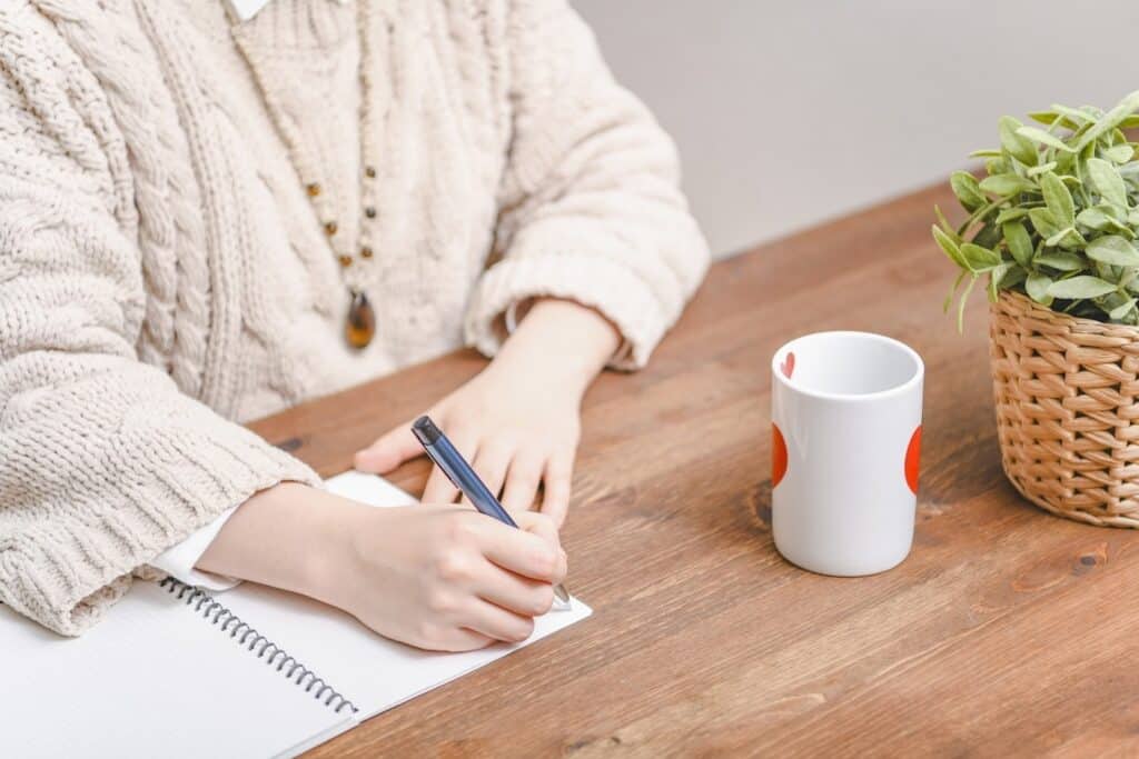Close-up of a person writing in a notebook on a wooden table, with a heart-patterned mug and a potted plant nearby — a calm and cozy workspace scene: Mindfulness Journal