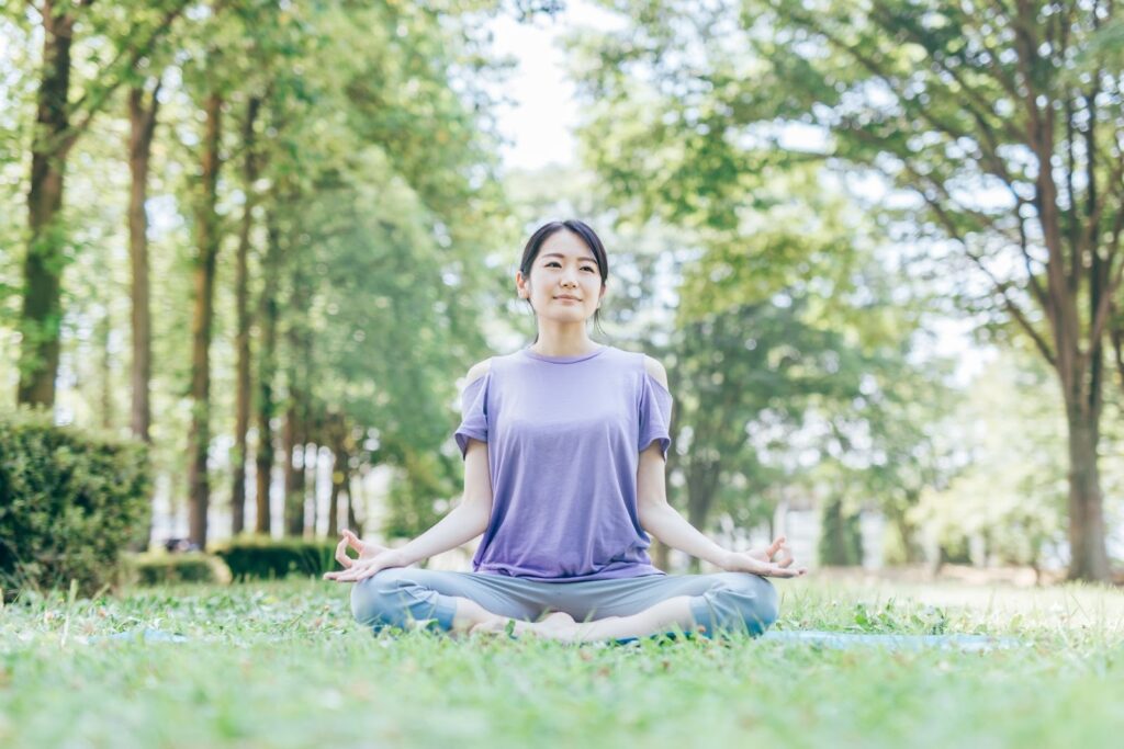 公園の芝生の上であぐらをかき、瞑想する女性。緑の木々に囲まれ、穏やかな表情で心を落ち着けている様子：スピリチュアル・プラクティス