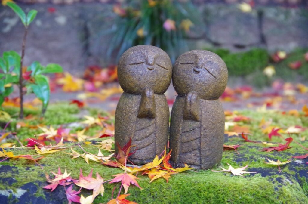 苔むした石の上に並ぶ、手を合わせた二体の小さなお地蔵様。周囲には紅葉が散り、秋の静けさと温かさを感じる情景：スピリチュアル・プラクティス