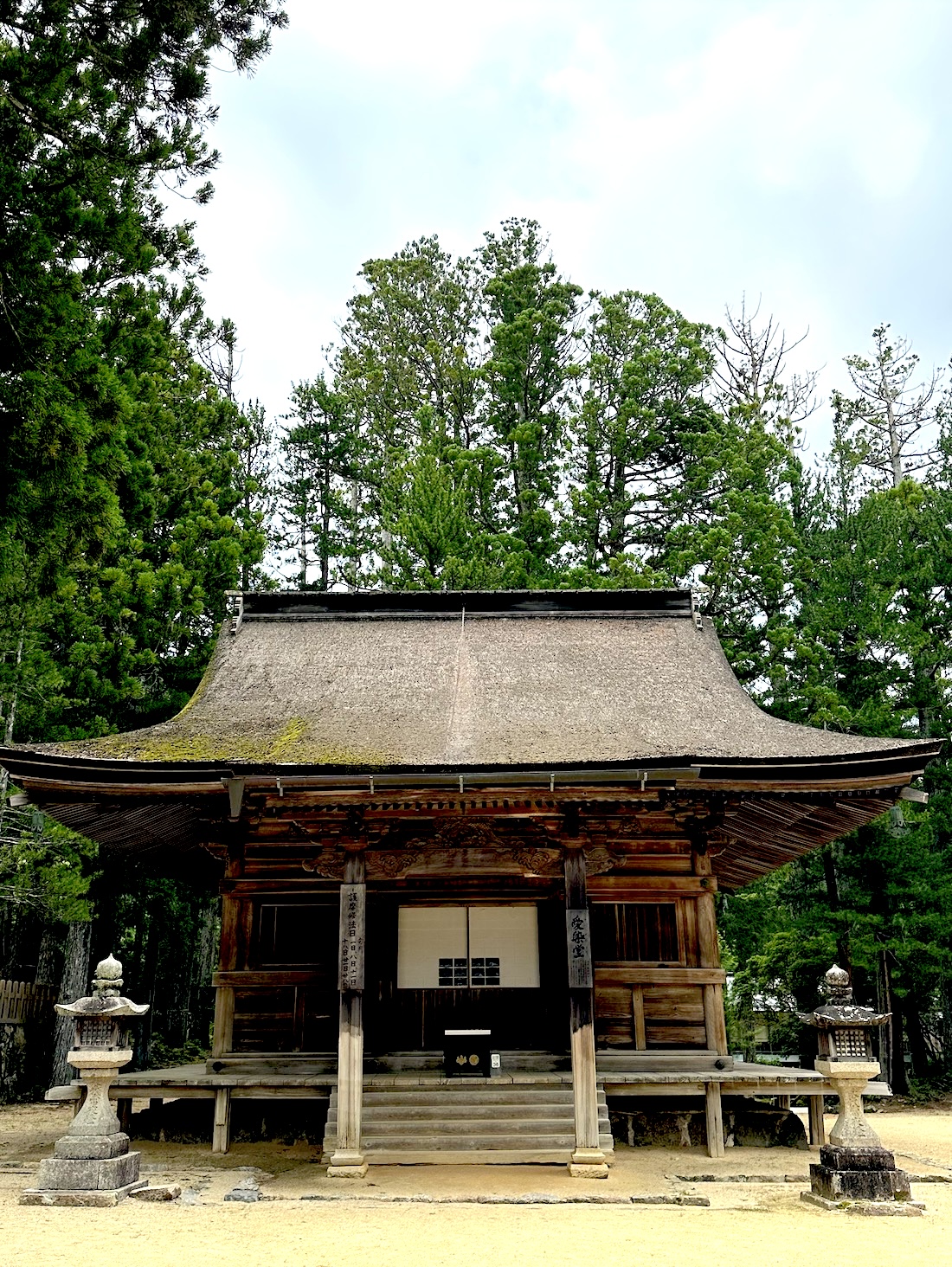 A front-facing view of a traditional Japanese temple hall with a thatched roof, wooden pillars, and an elevated veranda. Two stone lanterns stand on either side of the entrance, and tall cedar trees form a dense green backdrop behind the building: How to Meditate