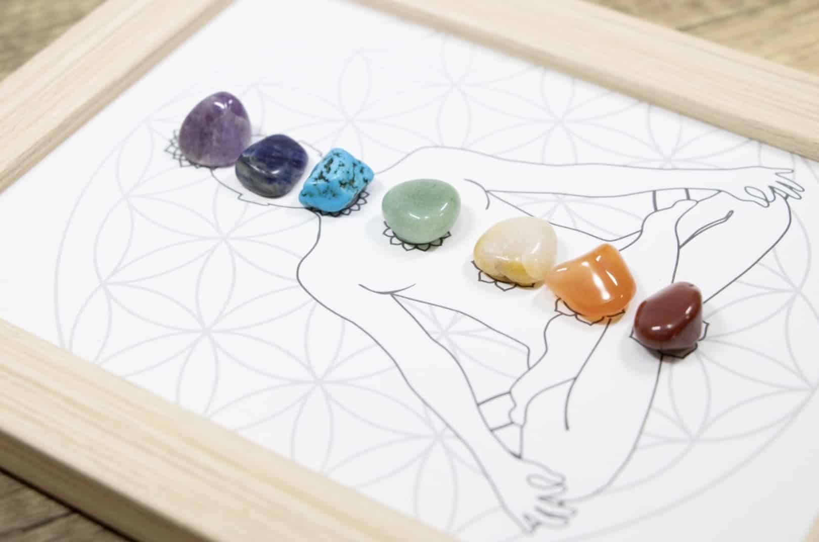 Several small polished stones of different colors are arranged in a line on a diagram showing a human silhouette, framed in wood: Chakra Meditation
