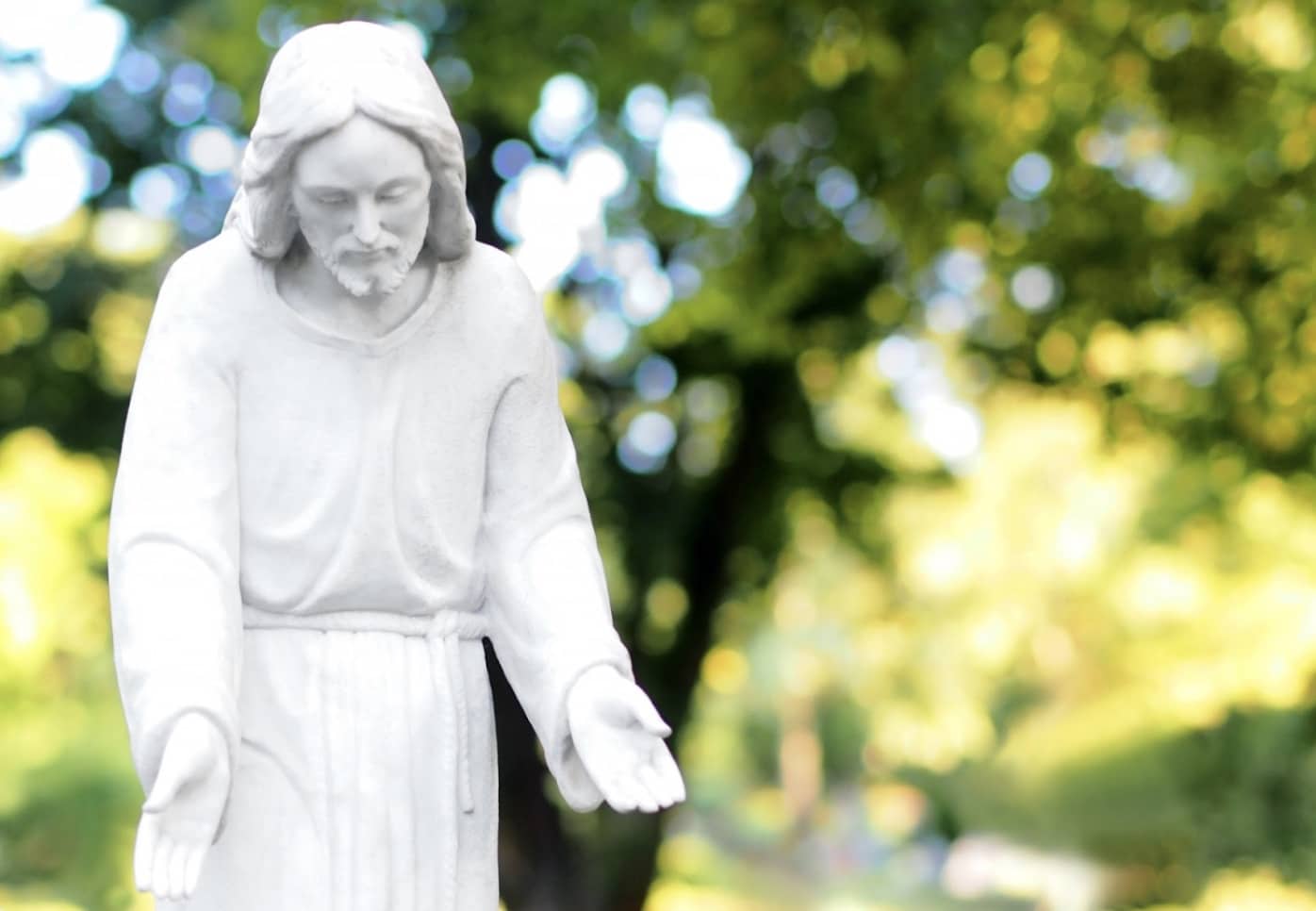 白いローブをまとい、両手を差し伸べる姿の石像。背景には柔らかな光が差し込む緑の木々が広がっている：クリスチャン・メディテーション