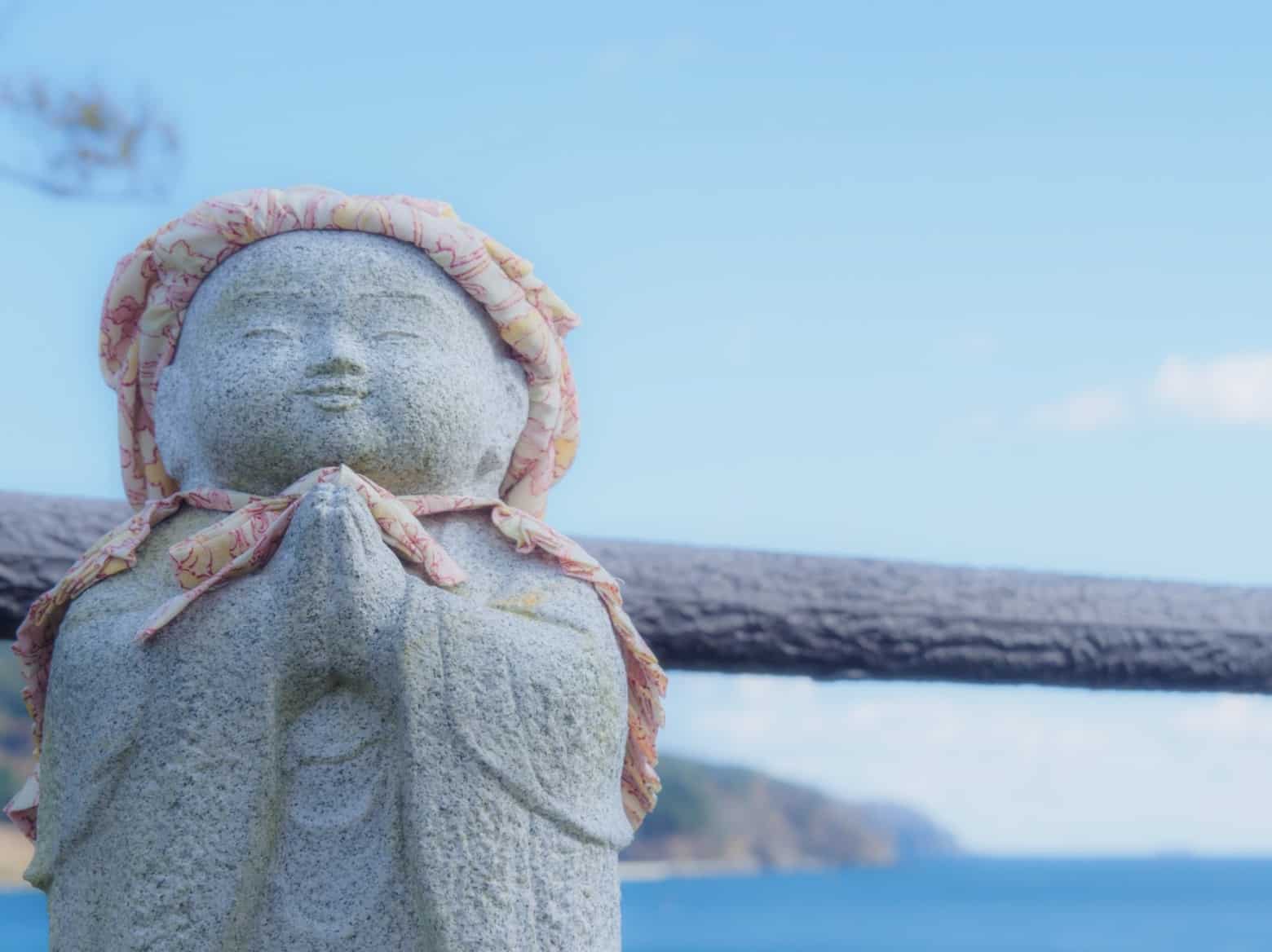 A childlike Jizo statue standing with hands together in prayer, set against a bright blue seaside background and wrapped in a patterned cloth: Mindful Souls