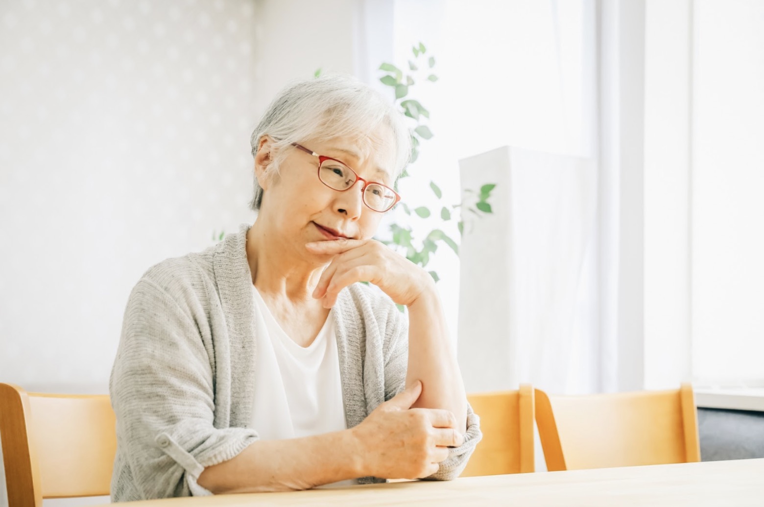 高齢の女性がテーブルに座り、顎に手を添えて物思いにふけっている：不安