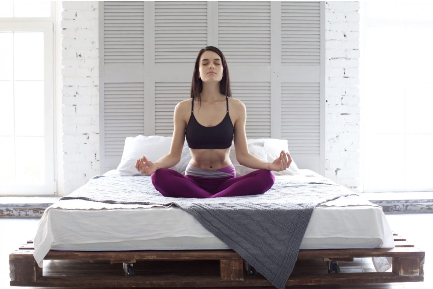 A person sitting cross-legged on a bed in a bright bedroom, eyes closed, with an upright posture and hands resting on their knees: Sleep Meditation