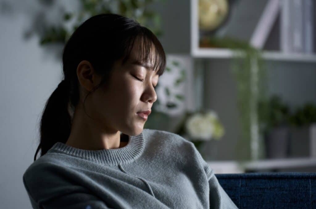 室内のソファに座り、目を閉じて静かに過ごす人物。背景には植物や棚があり、落ち着いた雰囲気の空間が広がっている：睡眠瞑想