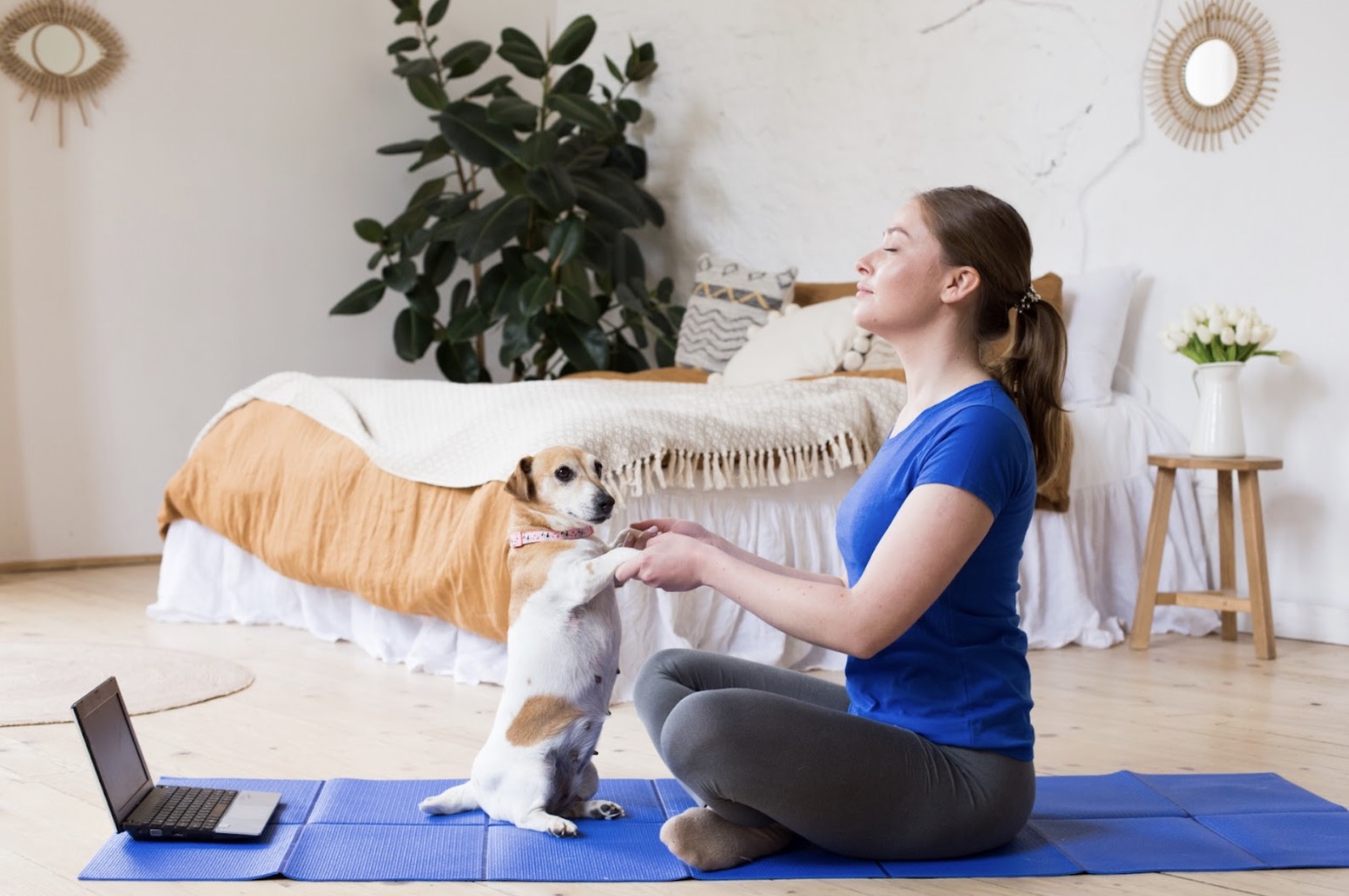 A woman sitting on a yoga mat indoors with her eyes closed, practicing meditation while gently holding paws with a small dog in front of her. A laptop is placed nearby in a calm bedroom setting: Types of Meditation