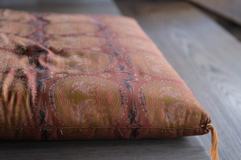 伝統的な文様が織り込まれた座蒲が木の台の上に置かれている。温かみのある色合いと質感が落ち着いた雰囲気を生み出している:座布団