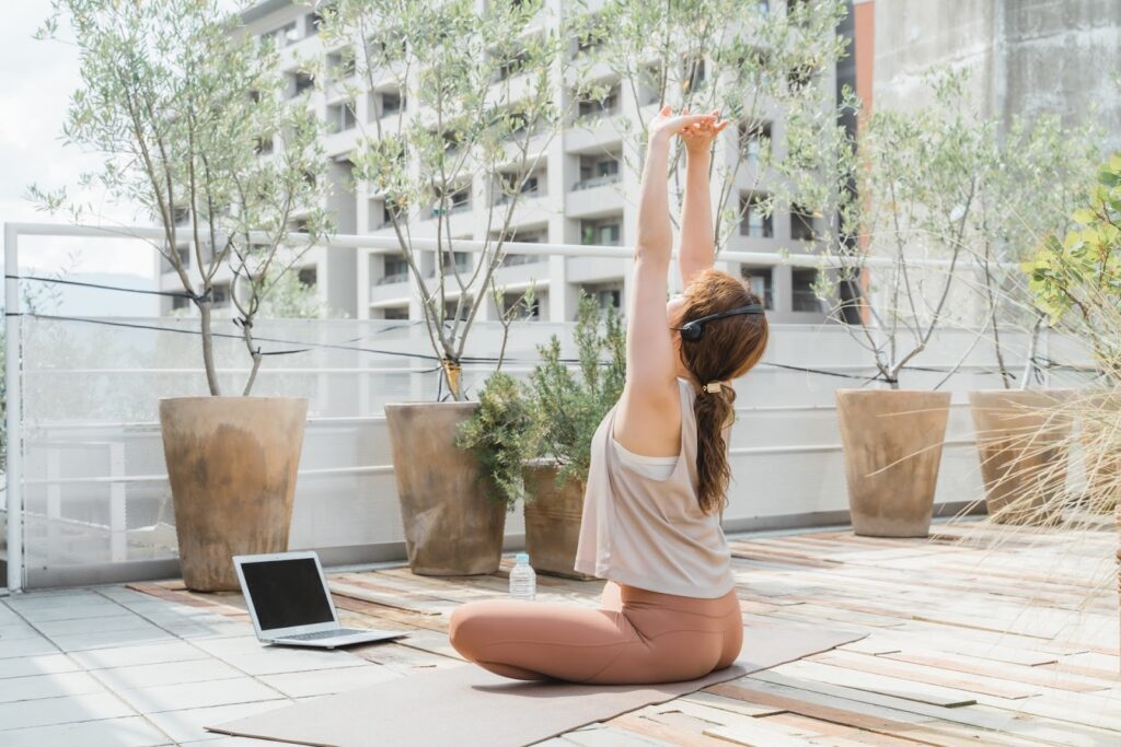 屋外テラスで座り、腕を伸ばしてストレッチをする人物。近くにはノートパソコンと水のボトルが置かれ、ヘッドセットを着用してオンラインレッスンを受けている様子:毎日の瞑想