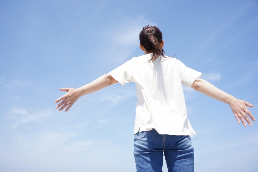 青空の下、腕を広げて立つ人物の後ろ姿。広がる空に向かって深く息を吸い込むような、開放的な雰囲気が伝わる:毎日の瞑想