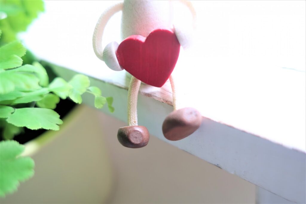 窓辺に座る小さな人形が赤いハートを抱えており、そばには緑の葉が見えている:慈愛の瞑想