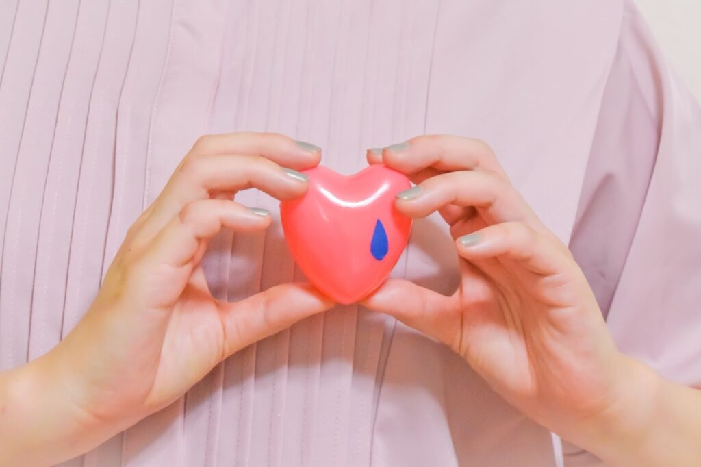 ピンク色の服を着た人が、青い涙のマークがついたハート型の小物を両手で持っている:慈愛の瞑想