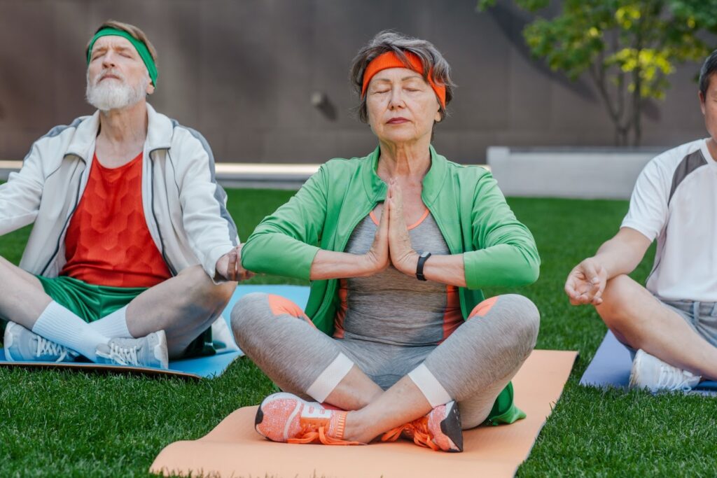 Older adults sit on yoga mats outdoors on the grass, eyes closed, practicing a meditation pose: Meditation Groups