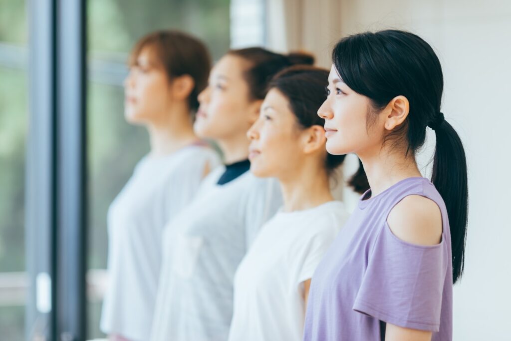 スタジオで横一列に立つ女性たちが、まっすぐ前を見つめて姿勢を整えている：瞑想コミュニティ