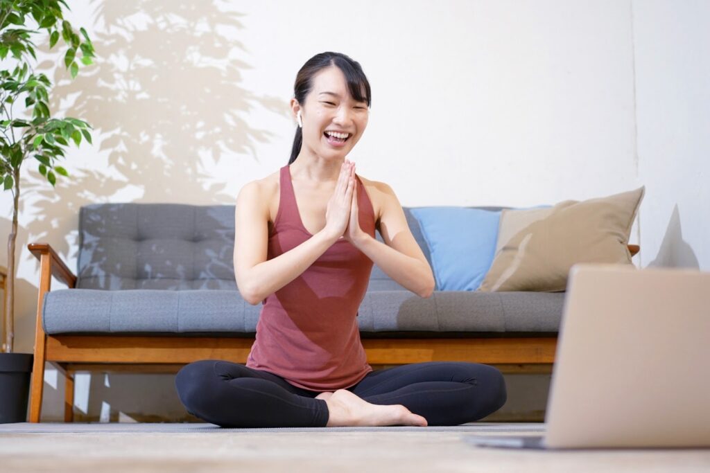 ヨガウェアを着た女性がリビングで座ってオンラインレッスンに参加し、手を合わせて笑顔であいさつしている：瞑想コミュニティ