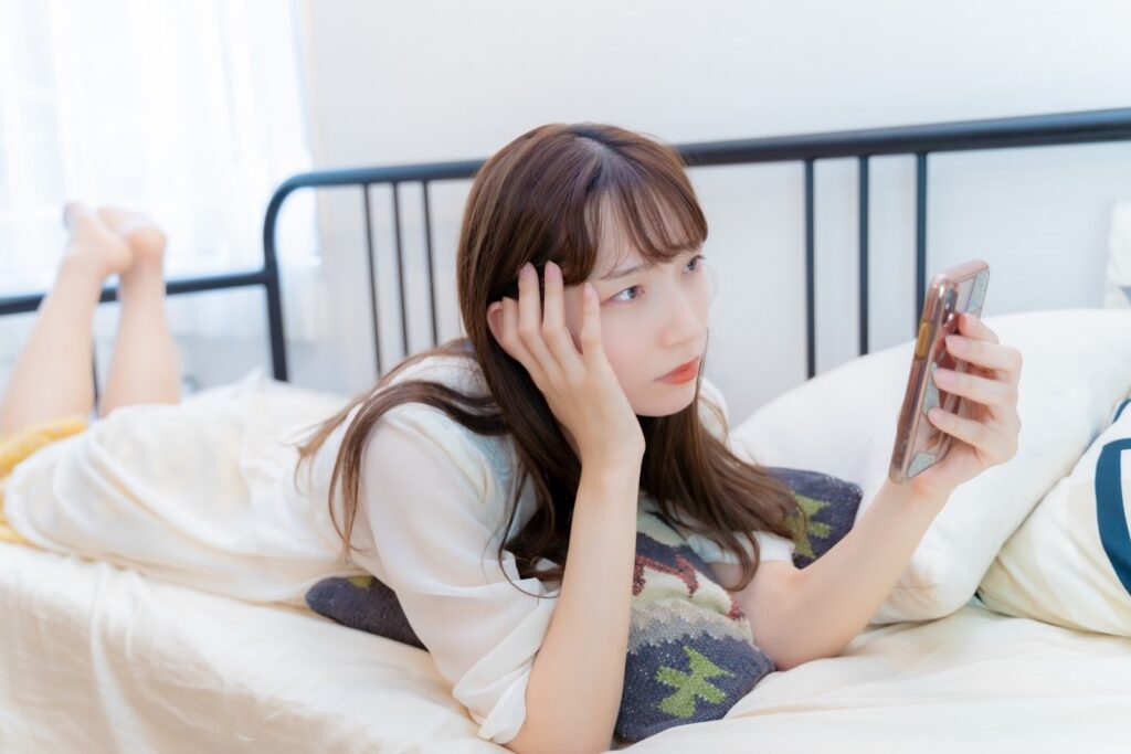 ベッドに横たわり、スマートフォンを手に持ち、こめかみに手を添えて物思いにふける女性：不安