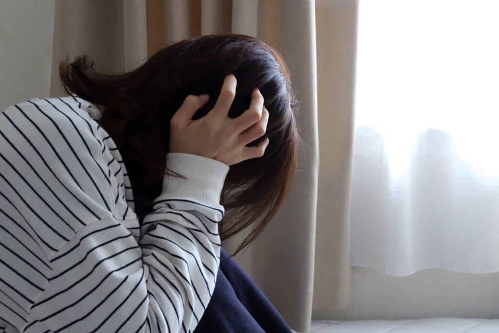 カーテン越しの光が入る室内で、膝を抱え、片手で頭を押さえながらうつむいて座る人物。静かな空間で考え込んでいる様子：瞑想の種類