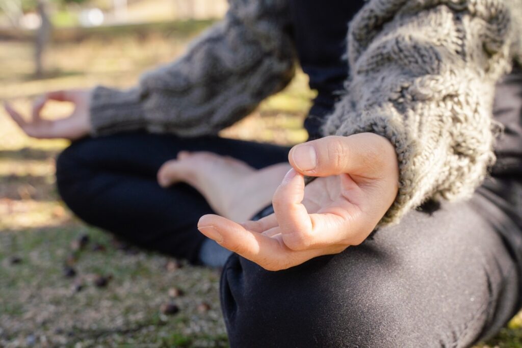 A person sitting cross-legged outdoors, with one hand resting palm-up and fingers gently curved, in natural daylight: Types of Meditation