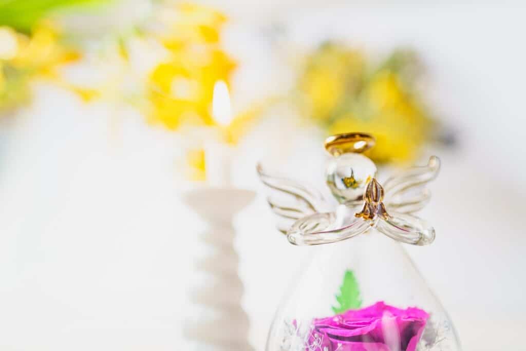 ガラス製の天使のオブジェと、その内部に飾られたピンクの花。背後には柔らかな光と黄色い花がぼんやりと映り、温かく優しい雰囲気をつくっている：クリスチャン・メディテーション