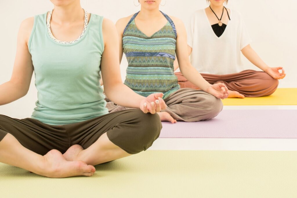 ヨガマットの上で足を組み、背筋を伸ばして座る複数の人物。手は膝の上に置かれ、静かな姿勢で呼吸に集中している様子：瞑想の種類