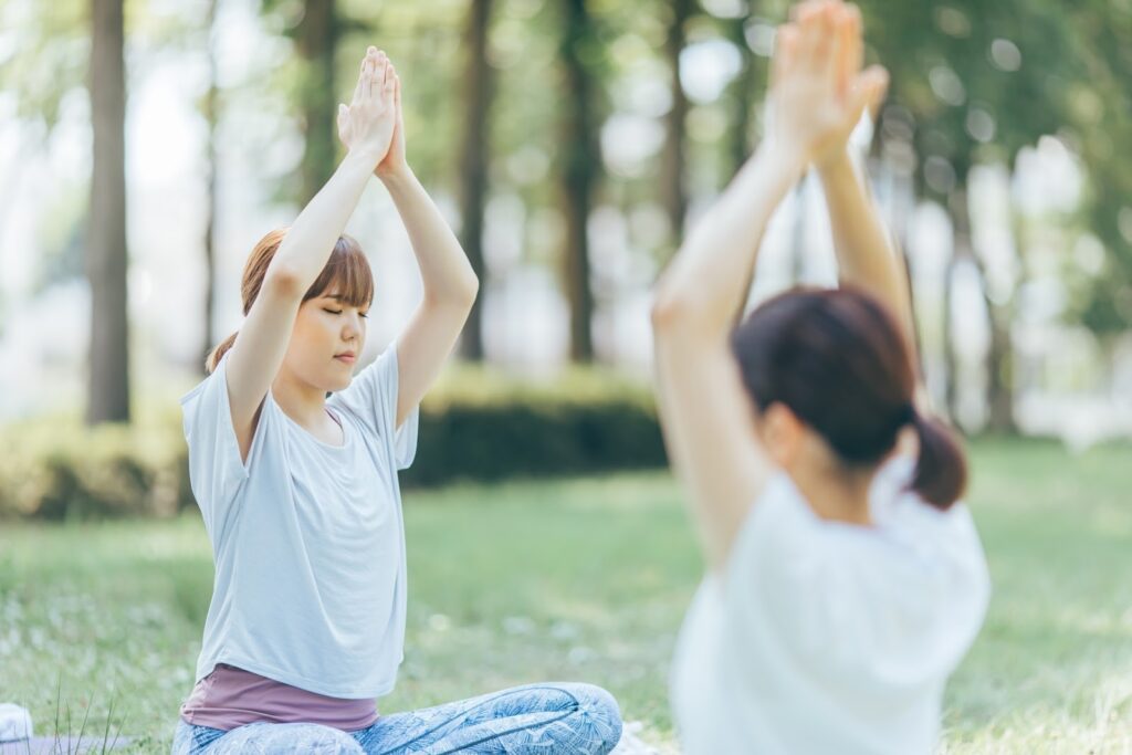 屋外の芝生で座り、両手を頭上で合わせてヨガのポーズをとる二人の人物。静かな森の中で深い呼吸に集中している様子：クリスチャン・メディテーション