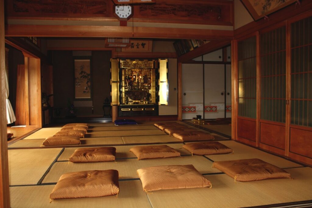 畳の上に座布団が整然と並べられた寺院の本堂。正面には金色の仏壇が置かれ、静寂と荘厳さが漂っている:座布団