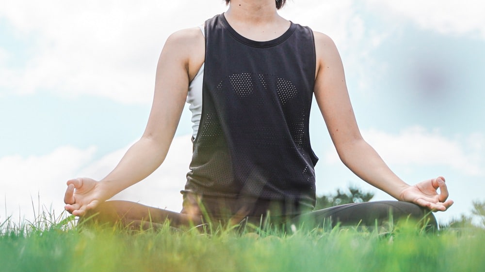 A person sitting cross-legged on the grass outdoors, meditating with relaxed open hands. Soft natural light enhances the calm atmosphere: Mindful Souls