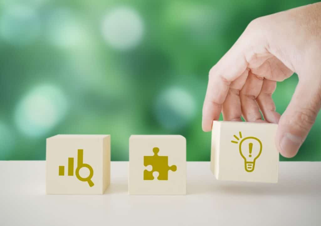 緑の背景の前で、手が電球アイコンの描かれた木製ブロックを並べて置いている。横にはグラフとパズルのアイコンが描かれたブロックがある：Mind Over Mood